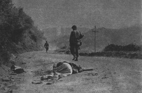 [Men of the 363rd Infantry advance toward a shell burst, passing a dead German in the road]