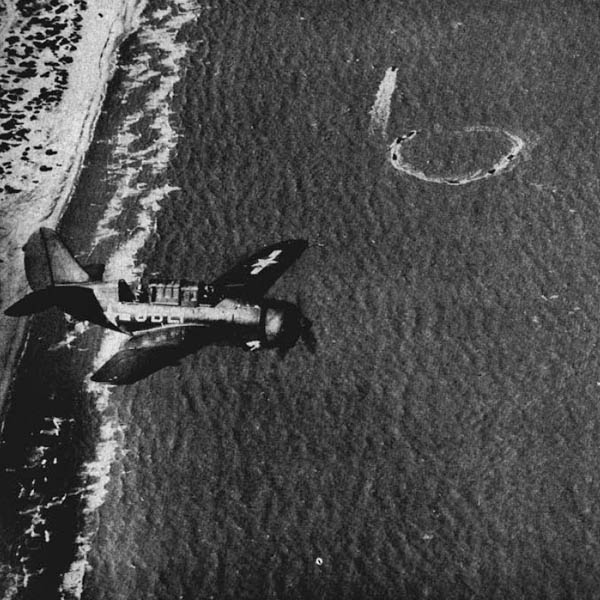 A Helldiver roars low over the shore as landing craft in maneuvers whirl through their buzzsaw circles before shooting toward the beach.