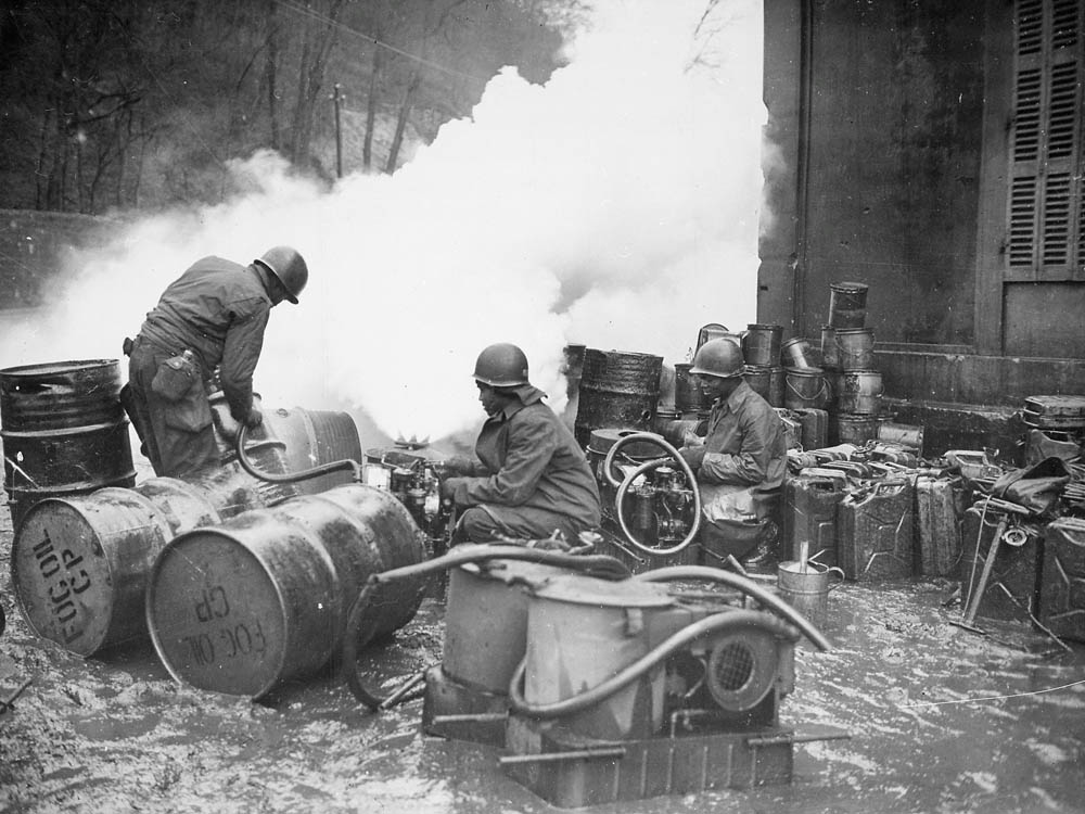 Soldiers of the 161st Chemical Smoke Generating Company, U.S. Third Army, move a barrel of oil in preparation to refilling an M-2 smoke generator, which spews forth a heavy cloud of white smoke.