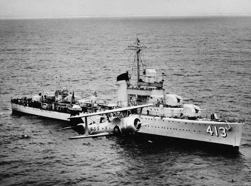 A Curtiss SBC-3 (Curtiss Model 77A) biplane from VS-6 of the USS Enterprise flies past USS Mustin (DD-413), a Sims-class destroyer of the United States Navy, during naval maneuvers in the Pacific in May 1940. (U.S. Navy Photograph.)