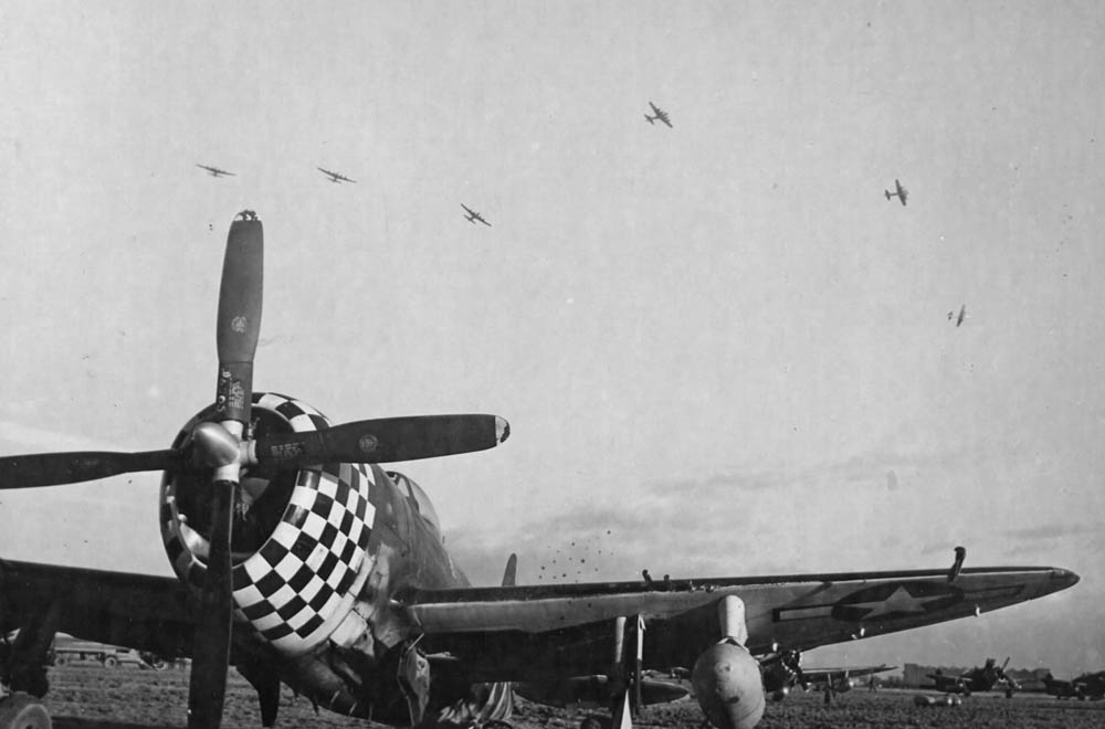 A group of Boeing B-17 Flying Fortress aircraft flies over a Republic P-47 Thunderbolt fighter on a field in England. (U.S. Air Force Photograph.)