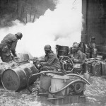 Soldiers of the 161st Chemical Smoke Generating Company, U.S. Third Army, move a barrel of oil in preparation to refilling an M-2 smoke generator, which spews forth a heavy cloud of white smoke.