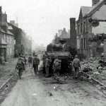 GI's advance past the wreck of an M4 Sherman tank of the 32nd Armored Regiment, 3rd Armored Division in Normandy. (U.S. Army Photograph.)