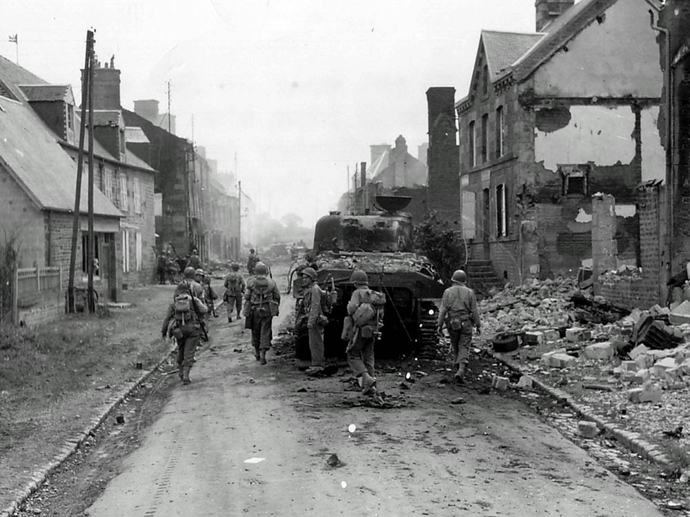 GI's advance past the wreck of an M4 Sherman tank of the 32nd Armored Regiment, 3rd Armored Division in Normandy. (U.S. Army Photograph.)